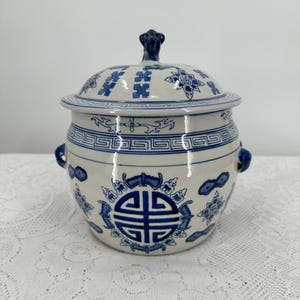 Peut inclure: Un pot en céramique blanc et bleu avec couvercle et motifs décoratifs. Le pot présente des poignées bleues, un bouton bleu et des motifs bleus et blancs. Le pot est de forme ronde et est probablement utilisé pour le stockage.