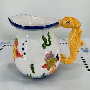 May include: White ceramic pitcher with a seahorse-shaped handle. The pitcher is decorated with colorful fish, seaweed, and bubbles. The rim is trimmed with blue. The handle is a golden yellow color.