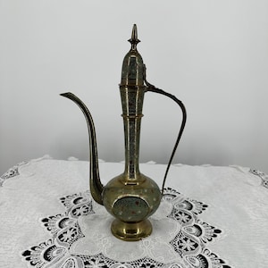 Puede incluir: Una jarra de agua antigua de latón con un pico largo y curvo y un asa. La jarra tiene intrincados patrones florales y geométricos en verde y rojo. La base y la parte superior son de un latón más oscuro.