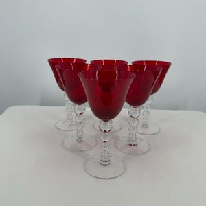 May include: A set of five wine glasses featuring deep red bowls and clear glass stems. Each stem is designed with a series of stacked spherical glass beads leading down to a circular clear glass base. The glasses are arranged in a triangular formation.