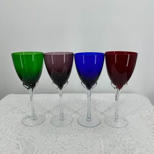 May include: Four colorful wine glasses with clear stems and bases. The glasses feature vibrant hues: green, purple, blue, and red. The glasses are arranged on a white lace tablecloth, creating a visually appealing display. These glasses are suitable for serving wine.