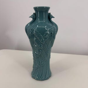 Könnte beinhalten: Eine türkisfarbene Keramikvase mit detailliertem Pfauenfedermuster. Die Vase hat einen ausgestellten Boden, einen langen Hals und zwei vogelartige Griffe. Die Vase ist etwa 25 cm hoch.