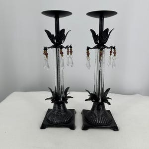 Puede incluir: Dos candelabros de metal negro con detalles de hojas y adornos colgantes de cristal y cuentas. Los candelabros tienen una base cuadrada y un diseño alto y delgado, ideal para añadir un toque de elegancia a cualquier espacio. Altura aproximada 30 cm.