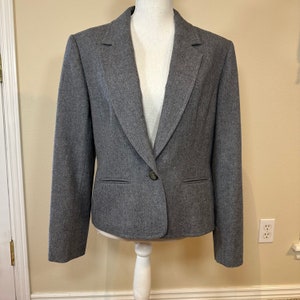 Puede incluir: Un blazer gris con cierre de un solo botón, dos bolsillos delanteros y solapa con muescas. La chaqueta se exhibe en un maniquí blanco y tiene un corte entallado. La tela parece ser un material tejido.