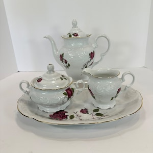 May include: A white porcelain tea set with a teapot, creamer, sugar bowl, and serving tray. The set is decorated with red roses and gold trim. The teapot has a lid and a curved spout. The sugar bowl has a lid and handles. The creamer has a handle.