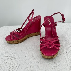 May include: Bright pink Prada wedge sandals with a glossy finish. The sandals feature a cork wedge heel and multiple straps across the foot. The ankle strap has a silver buckle. The Prada logo is visible on the insole.