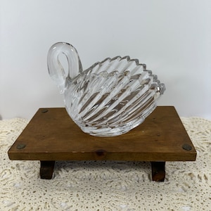 以下が含まれることがあります： テクスチャ加工された渦巻き模様の、透明なガラス製の白鳥型皿。白鳥は湾曲した首と幅広の開いたボウルを持っています。短い脚の付いた、小さな長方形の木製スタンドの上に置かれています。