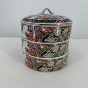May include: A three-tiered ceramic food container with a matching lid, featuring a detailed pattern of orange, yellow, and white flowers against a dark blue background with gold trim accents on the edges of each circular section.