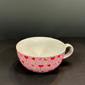 Puede incluir: Un cuenco de cerámica rosa con interior blanco y asa. El exterior está decorado con un patrón de corazones rojos y rosas sobre tallos verdes. Ideal para servir sopas o cereales. Diámetro aproximado de 15 cm.
