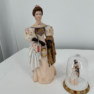 May include: A porcelain figurine of a woman in a long, cream-colored dress with floral accents, holding a shawl and a small purse. A smaller figurine of a couple under a glass dome is also present. The woman's dress has rose details.