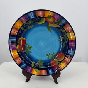 May include: A decorative ceramic plate with a blue center and a colorful striped border. The plate features painted illustrations of tomatoes, peppers, and olives with green leaves. The plate is displayed on a wooden stand.