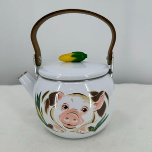 May include: White enamel teapot with a pig illustration on the front. The teapot has a brown handle and a corn-shaped lid handle. The pig has brown spots and a pink snout. Green and yellow corn is on the lid.