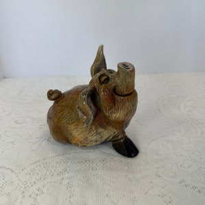 May include: A ceramic pig figurine in shades of brown and tan, with a glossy finish. The pig is depicted in a playful pose, with its snout pointing upwards and one ear raised. The figurine has black hooves and a curly tail.