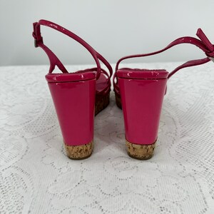 May include: Pair of bright pink wedge sandals with a glossy finish. The sandals have a cork sole and a thin ankle strap. The wedge heel provides height and support. The shoes are designed for a stylish look.