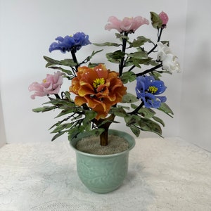 Pode incluir: Um arranjo floral decorativo de vidro num vaso de cerâmica verde claro. O arranjo apresenta flores de vidro coloridas em tons de laranja, azul, rosa e branco, com folhas de vidro verde. O vaso tem um design em relevo detalhado e é preenchido com pequenas pedras brancas.