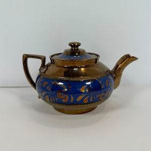 May include: An antique teapot with a bronze finish and a blue band with gold floral patterns. The teapot has a matching lid and handle, and a curved spout. The design is ornate and elegant. The teapot is likely made of ceramic.