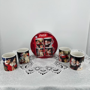 Puede incluir: Un juego de seis tazas navideñas de Coca-Cola de cerámica. Las tazas presentan ilustraciones de Papá Noel en rojo, blanco y negro. Las tazas están dispuestas sobre un mantel de encaje blanco, con una lata redonda roja que contiene cuatro tazas adicionales.
