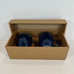 May include: Two navy blue ceramic mugs with handles are presented inside a brown cardboard gift box. The mugs have a glossy finish and a dark blue color. The box is open, revealing the mugs, which are side by side.