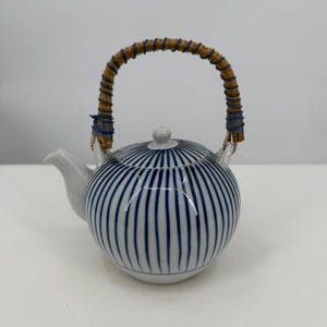 Può includere: Teiera in ceramica bianca con motivo a righe blu e bianche. La teiera ha un beccuccio curvo e un manico intrecciato con accenti blu. Il coperchio ha un piccolo pomello bianco. Un oggetto decorativo per gli amanti del tè.