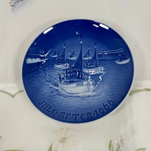 May include: A round, blue and white decorative plate featuring a nautical scene with several sailboats on the water. The plate has the text "JULE-AFTEN 1966" in white lettering around the bottom edge. The plate is on a white surface.