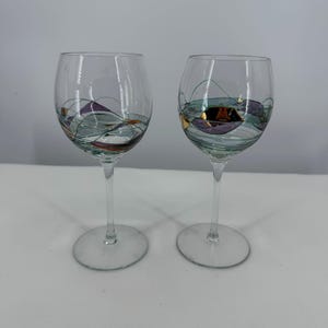 May include: Two clear glass wine glasses featuring a decorative painted design on the bowls. The artwork consists of thin black lines forming abstract shapes, accented with patches of metallic gold, deep purple, and soft teal paint colors.