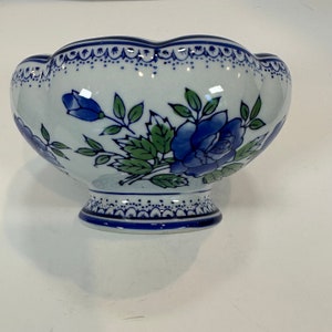 May include: A white ceramic bowl with a scalloped rim, decorated with blue roses and green leaves. The bowl has a pedestal base and blue trim. The floral design is hand-painted.