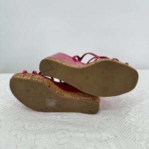 May include: Pair of bright pink wedge sandals with cork soles and thin straps. The sandals have a glossy finish and are designed with a wedge heel. The soles show signs of wear. The sandals are a size 37 1/2.