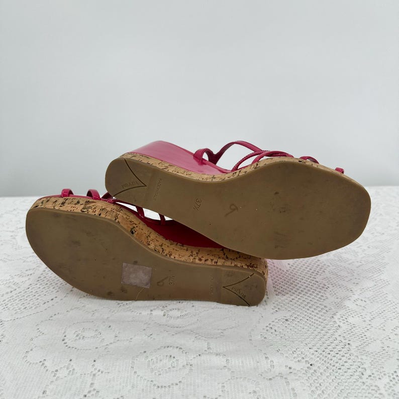 May include: Pair of bright pink wedge sandals with cork soles and thin straps. The sandals have a glossy finish and are designed with a wedge heel. The soles show signs of wear. The sandals are a size 37 1/2.