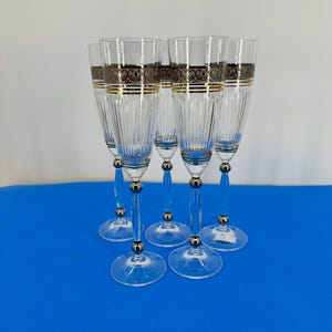 Peut inclure: Ensemble de six flûtes à champagne transparentes avec des bandes décoratives dorées et noires près du bord. Les tiges ont un détail de boule argentée et une base transparente. Les flûtes sont disposées sur une surface bleue.