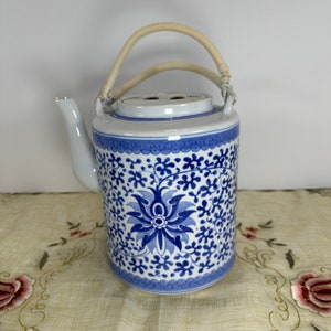 May include: A blue and white ceramic teapot with a floral pattern. The teapot has a cylindrical body, a curved spout, and a woven handle. The blue floral design covers the body of the teapot.