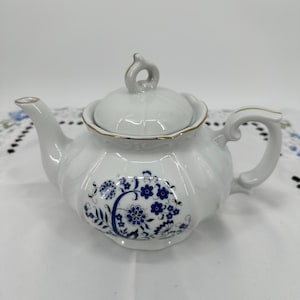 Op de afbeelding: Witte keramische theepot met deksel en handvat. De theepot heeft een blauw bloemmotief en gouden rand. De tuit en het handvat zijn gebogen. Het deksel heeft een kleine lusgreep. De theepot staat op een wit oppervlak.