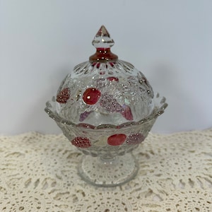 Puede incluir: Un cuenco de cristal transparente para caramelos con tapa, con un diseño de frutas con detalles en rojo. El cuenco tiene una base de pedestal y un borde festoneado. La tapa está rematada con un remate de punta roja.