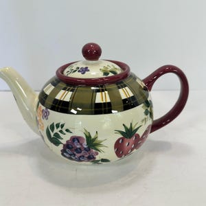 Op de afbeelding: Een keramische theepot met een crèmekleurige basis, versierd met een groen en zwart ruitpatroon. De theepot heeft een bordeauxrode handgreep, deksel en rand, en is versierd met geschilderde vruchten en bloemen.
