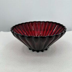 May include: A ruby red glass bowl with a fluted design. The bowl has a scalloped rim and sits on a small, star-shaped base. The bowl's interior is a vibrant red, while the exterior is a deep, dark red.