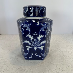 May include: A dark blue ceramic jar with a lid. The jar has a hexagonal shape and is adorned with white floral and leaf patterns. The lid mirrors the design. The jar is probably for storage or decorative purposes.