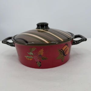 Puede incluir: Una cazuela redonda de cerámica con tapa, que presenta una base de color rojo intenso decorada con hojas y frutas pintadas. La tapa tiene un acabado brillante con rayas marrones, verdes y crema. Incluye dos asas laterales negras.
