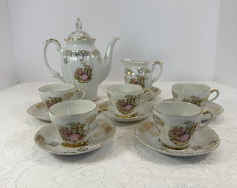 May include: A white porcelain tea set with gold trim. The set includes a teapot, creamer, sugar bowl, and six teacups with saucers. Each piece features a decorative scene with figures in a garden setting.