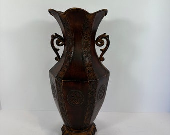 May include: A decorative brown vase with a unique shape and ornate detailing. The vase features two curved handles and embossed patterns. The base has a decorative, scalloped design. The vase is approximately 14 inches tall.