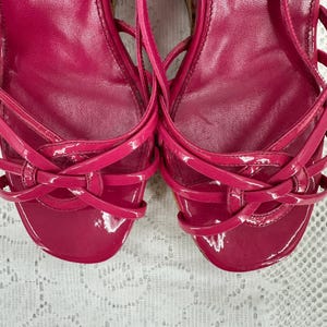 May include: Pair of bright pink, strappy sandals with a glossy finish. The sandals feature multiple thin straps that crisscross over the foot and ankle. The design includes a rounded toe and a low heel, perfect for a summer look.