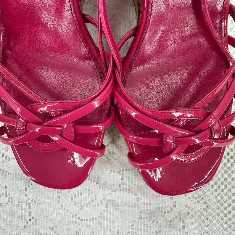 May include: Pair of bright pink, strappy sandals with a glossy finish. The sandals feature multiple thin straps that crisscross over the foot and ankle. The design includes a rounded toe and a low heel, perfect for a summer look.