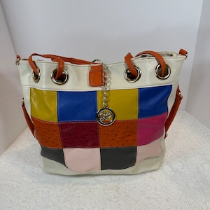 May include: A patchwork tote bag with a variety of colors including white, yellow, blue, pink, and orange. The bag has a silver charm and orange straps. The bag is made of leather.