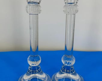 May include: A pair of clear glass candle holders. Each has a circular base with a ridged design, a long, thin stem, and a cup-shaped top. The candle holders are made of clear glass and are designed to hold candles.