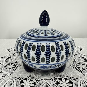 May include: A white and blue ceramic incense burner with a lid and three feet. The burner has a pierced design with blue leaf patterns and a dark blue finial on the lid. The shape is round.