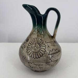 May include: A ceramic pitcher with a bulbous body and a curved handle. The top section features a dark green glaze that fades into a light grey, textured base. The surface is decorated with black line drawings of a sun, animals, and geometric shapes.
