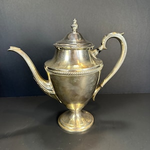 May include: A silver-colored teapot with a decorative handle and spout. The teapot has a lid with a small finial and a rope-like design around the middle. The teapot sits on a pedestal base.