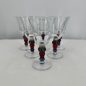 May include: Six clear wine glasses with decorative stems. The stems have red, blue, and gold accents. The glasses have a classic tulip shape and sit on a clear, round base. The glasses are arranged on a white surface.