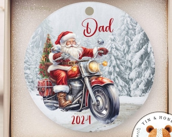 Puede incluir: Adorno navideño redondo con un Papá Noel de dibujos animados conduciendo una motocicleta a través de un bosque nevado. El adorno es blanco con detalles rojos y negros. El texto "Dad" y "2024" están impresos en el adorno.