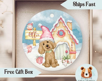 May include: A personalized Christmas ornament featuring a brown dog wearing a pink Santa hat in front of a gingerbread house with the name "Ollie" on a sign. The ornament is on a white background with a blue sky and snow.