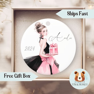 May include: A white round ornament with a black and pink illustration of a woman holding a pink gift box. The woman has blonde hair and is wearing a black dress with a pink bow. The text "2024" and "Amelia" are written on the ornament.