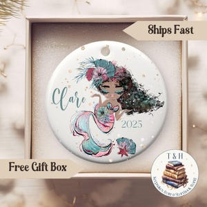 May include: A white ceramic ornament featuring a mermaid illustration with teal, pink, and black accents. The ornament has the name "Clare" and the year "2025" printed on it. The ornament is in a gift box.
